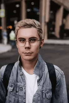 a young man wearing glasses and a denim jacket is standing on a street