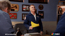 a man in a blue jacket is holding a yellow piece of paper with the nbc logo on the bottom right