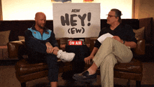 two men sit on a couch in front of a sign that says " hey "