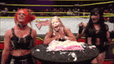 three women are standing around a table with a cake on it in front of a wrestling ring with the letter w on it
