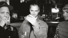 three men are standing next to each other in a black and white photo and one of them is smoking a cigarette .