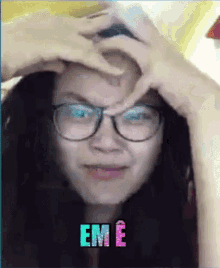 a woman wearing glasses is making a heart shape with her hands on her forehead with the word em e written on the bottom