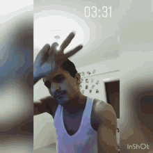 a man wearing a white tank top is waving his hand in front of a wall with pictures on it