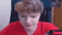a young man in a red shirt is sitting in a chair and looking at the camera .