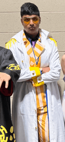 a man wearing a white coat and sunglasses is standing with his arms crossed .