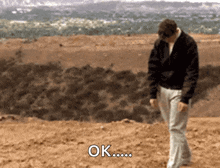 a man in a black jacket is walking on a dirt road and says ok ..