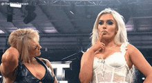 two women are standing next to each other in a wrestling ring and one is holding her finger to her mouth .
