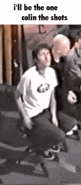 a man in a white shirt is sitting on a chair with a caption that says i 'll be the one colin