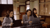 three women are sitting at a table using laptops and tablets .
