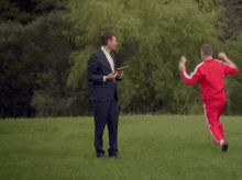 a man in a suit holds a tablet while a man in red jumps in the air with his arms in the air