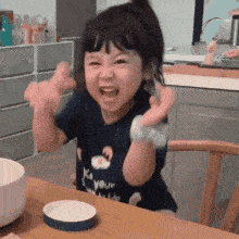 a little girl is sitting at a table with her arms in the air and making a funny face .