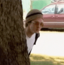 a man is peeking out from behind a tree with a car in the background .