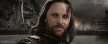 a man with long hair and a beard is looking at the camera with the words `` for kuda '' written on his face .