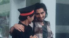 a man wearing a black hat with a red stripe is hugging a man wearing a plaid shirt