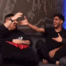 two men are sitting on a couch laughing and one has a shirt that says ' tg ' on it