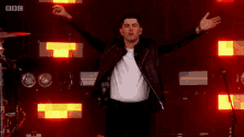 a man in a leather jacket is standing on a stage with his arms outstretched and a bbc logo in the background