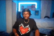 a man wearing a black shirt with a red heart on it is sitting on a bed in a room .