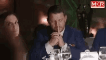 a man in a suit is sitting at a table with his hands folded in front of his mouth and says " crap "