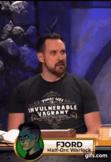 a man with a beard is sitting at a table wearing a black shirt that says `` invulnerable vagrant '' .