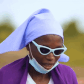a woman wearing sunglasses and a mask has a purple scarf on her head