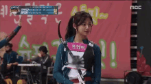 a woman holding a bow and arrow in front of a scoreboard that says mbc on it