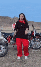 a woman in red pants and a black shirt is standing in front of a motorcycle .