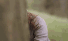 a woman wearing a hooded jacket and a hat is hiding her face behind a tree