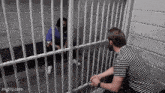 a man in a striped shirt is sitting in a jail cell talking to a man behind bars .
