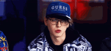 a young man wearing a guess hat and glasses is sitting down .