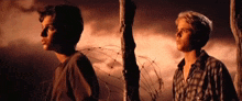 two young men are standing next to each other in front of barbed wire .