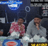 two men are sitting at a table with boxes of pizza and a clock that reads 14:21:02