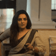 a woman is sitting on a couch holding a cell phone and the name sanjo_sebi is on the bottom of the picture