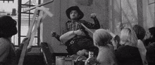 a man in a cowboy hat is playing a guitar in front of a group of people in a room .