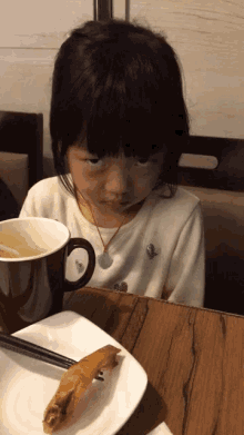 a little girl is sitting at a table with a cup of soup and a plate of food