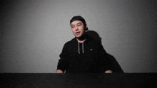 a man wearing a black hat and a black hoodie is standing in front of a white wall