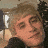 a young man with blonde hair is sitting in front of a christmas tree and smiling .