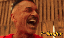 a man with a mustache is laughing in front of a sign that says ' the surfer '