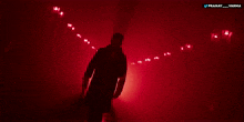 a man in a trench coat stands in a dark room with red lights behind him and the words pranay and varma below him