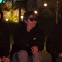 a man wearing sunglasses and a black hoodie is kneeling down in front of a group of people .