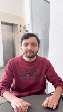 a man wearing a maroon ocean master sweatshirt sits at a desk