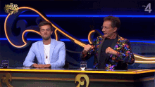 two men are sitting at a table with a masked singer logo on the wall behind them