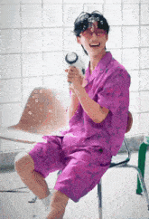 a man in a purple shirt and shorts is sitting on a chair holding a hair dryer and blowing bubbles .