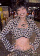 a woman wearing a plaid shirt and a crop top is standing on a street with her hands on her hips .