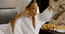 a woman in a bathrobe with a towel wrapped around her head is sitting on a bed eating food .