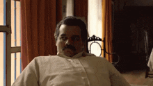 a man with a mustache is sitting in a chair in front of a window .