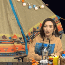 a woman is sitting in front of a teepee with a bottle of water and a box of cheetos
