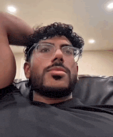 a man with curly hair and a beard wearing glasses is sitting on a couch .