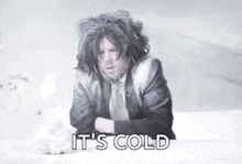 a man in a suit and tie is sitting on a bed with his arms crossed and says `` it 's cold '' .