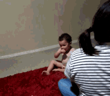 a baby is sitting on a red rug with a woman looking on