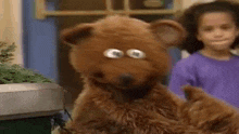a brown teddy bear with big eyes is standing next to a little girl .